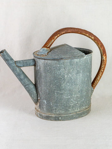 Antique French zinc oval watering can with weathered finish and rusted handle, on light fabric background.