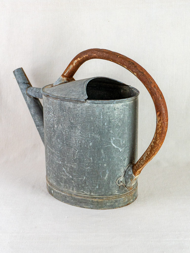 Antique French zinc oval watering can with weathered finish and rusty handle on light surface.