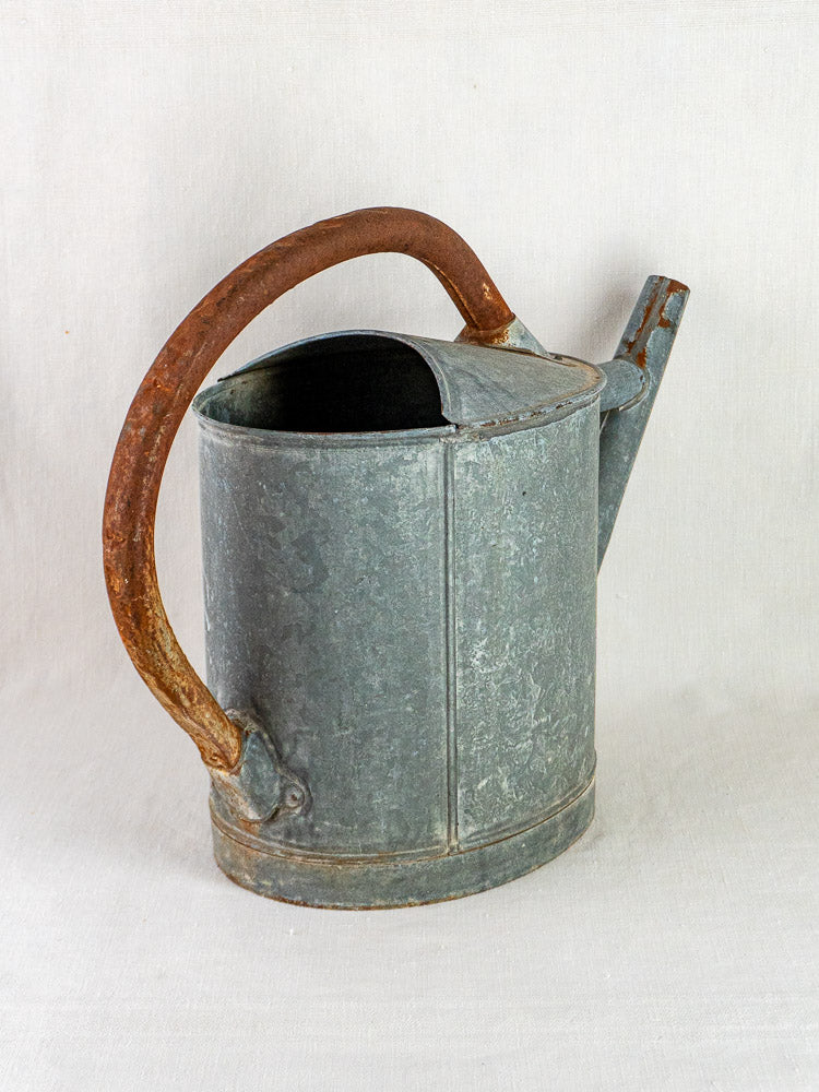 Antique French zinc oval watering can with rusted brown handle and straight spout on pale surface.