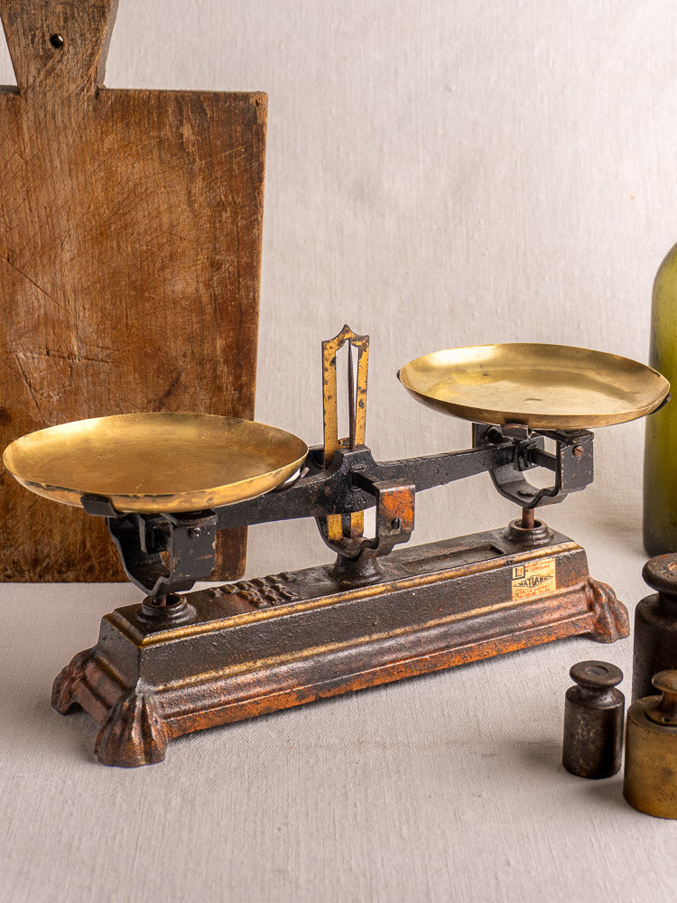 Antique French Roberval balance scale, cast iron with two brass pans on ornate black and brown base, light gray background.