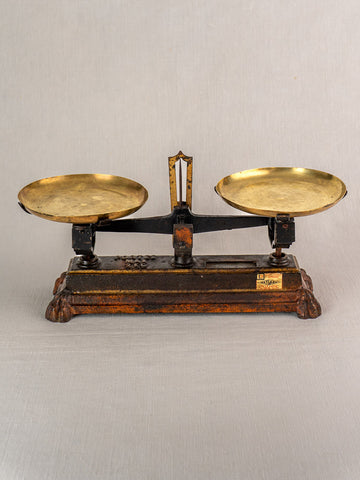 Antique French Roberval balance scale, cast iron with two brass pans on ornate black and brown base, light gray background.