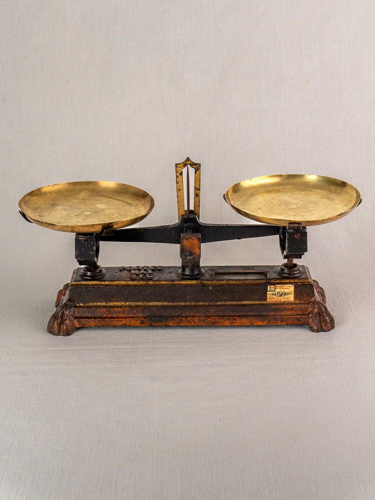 Antique French Roberval balance scale, cast iron with two brass pans on ornate black and brown base, light gray background.