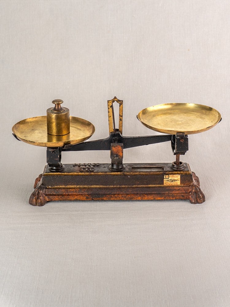 Antique French cast iron and brass balance scale with ornate base, one brass pan holding a metal weight, on light background.