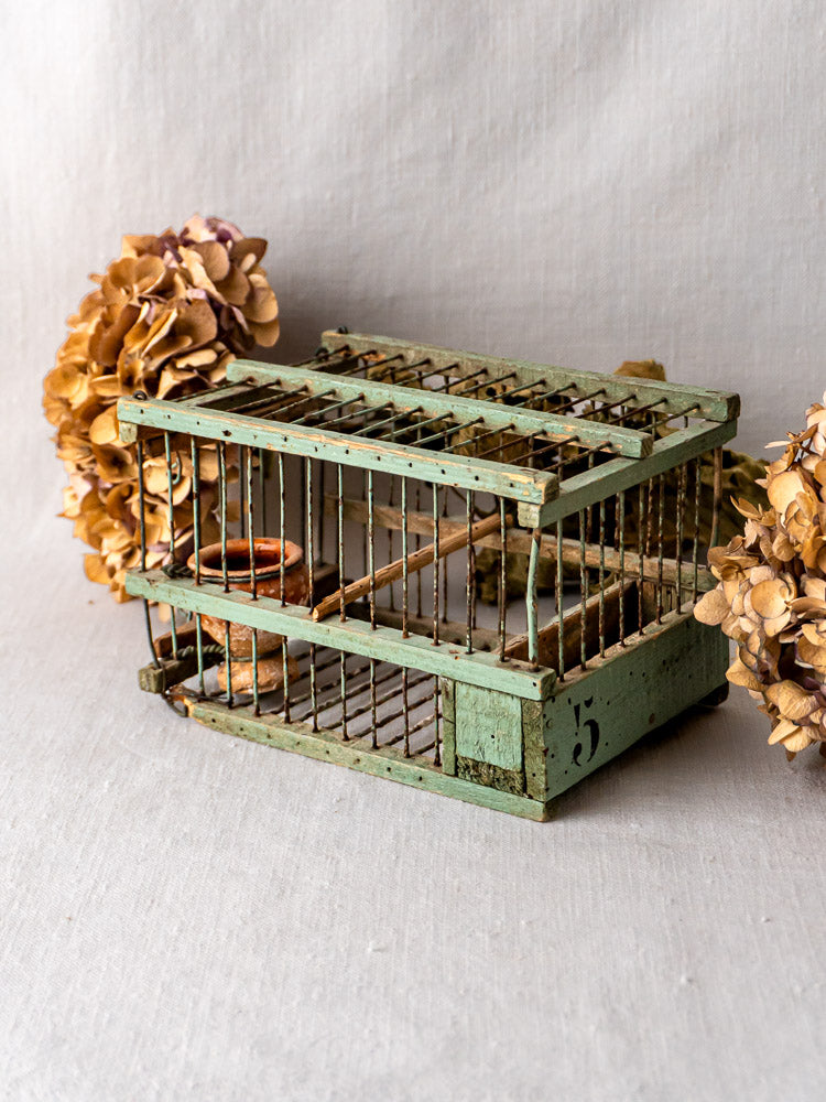 Antique French sage green decoy bird cage with number 5, displayed on light fabric, dried hydrangeas nearby.