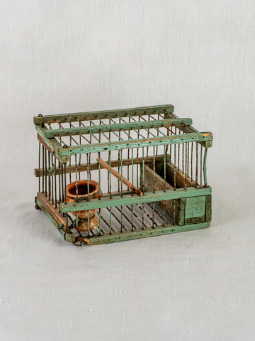 Antique French sage green decoy bird cage with wooden perch and clay water cup on neutral background.