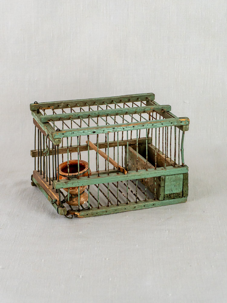 Antique French sage green decoy bird cage with wooden perch and clay water cup on neutral background.