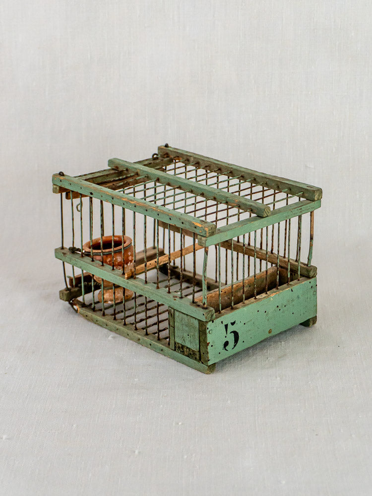 Antique French sage green metal birdcage with number 5, rusted bars, and clay water dish on fabric.