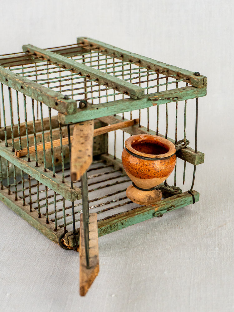 French sage green decoy bird cage with open door, wooden perch, and brown clay water pot.