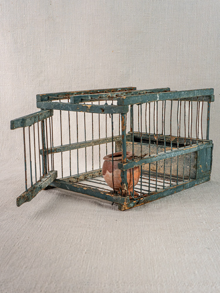 Late 19th-century Provençal bird cage with blue paint and terracotta cup, door open, on light fabric.