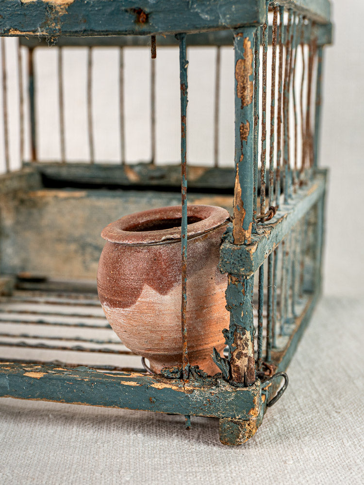 Provençal bird cage with terracotta cup, late 19th century blue paint, on neutral fabric surface.