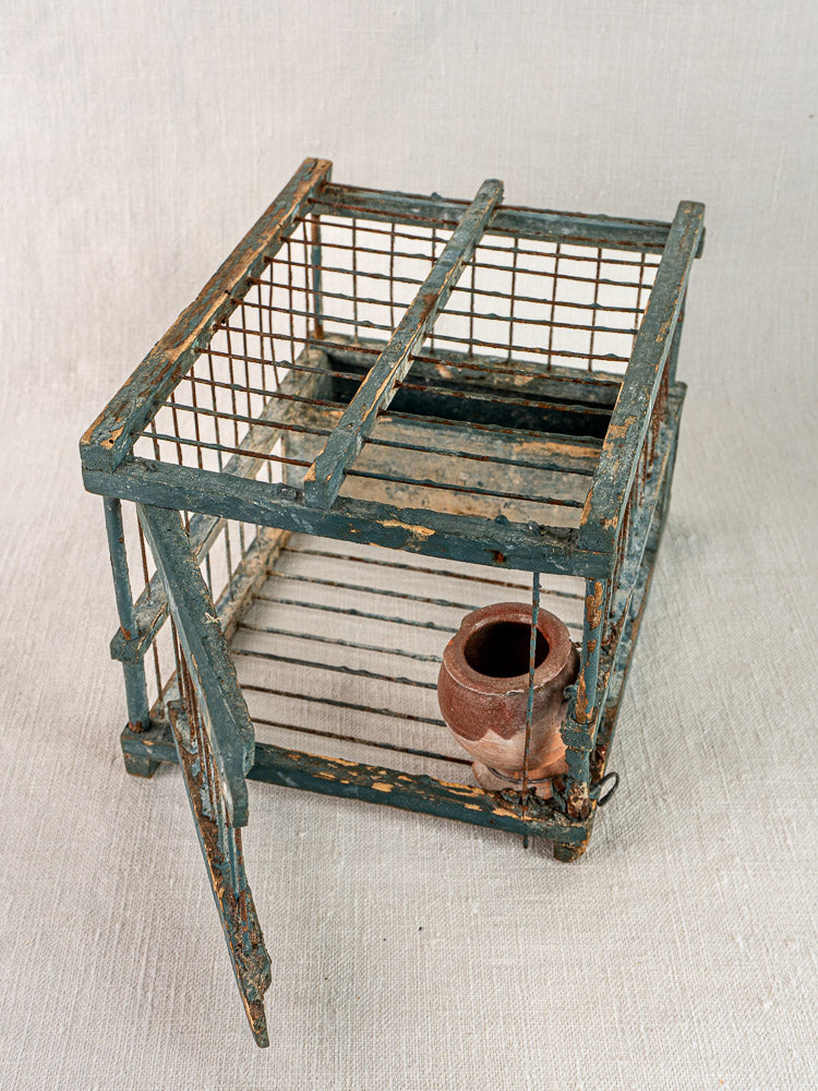 Antique Provençal bird cage with original blue paint and terracotta cup, displayed open on a light fabric surface.