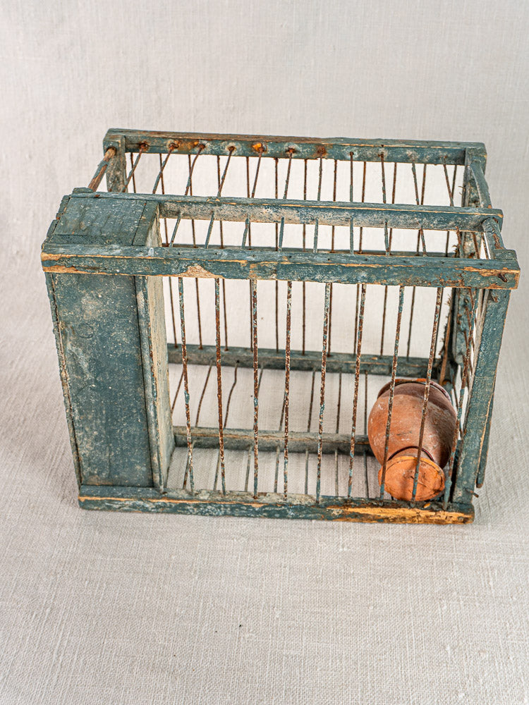 Late 19th-century Provençal bird cage with original blue paint and terracotta cup on light fabric surface.