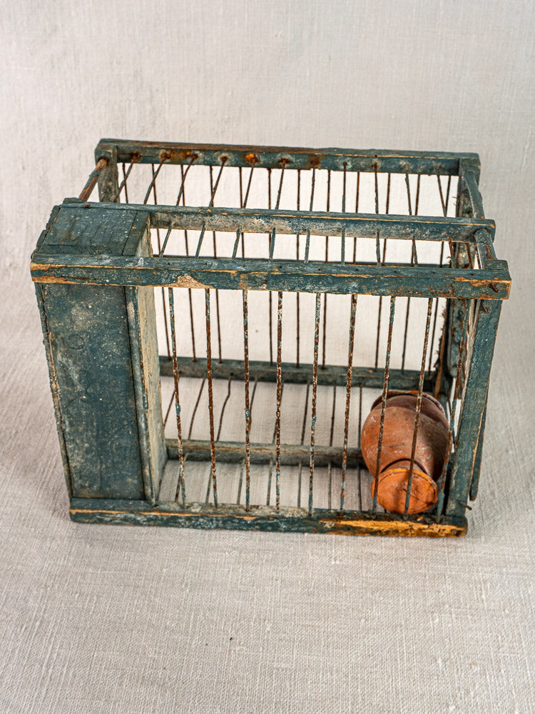 Antique French bird cage with original blue paint and terracotta cup, displayed on light grey fabric background.