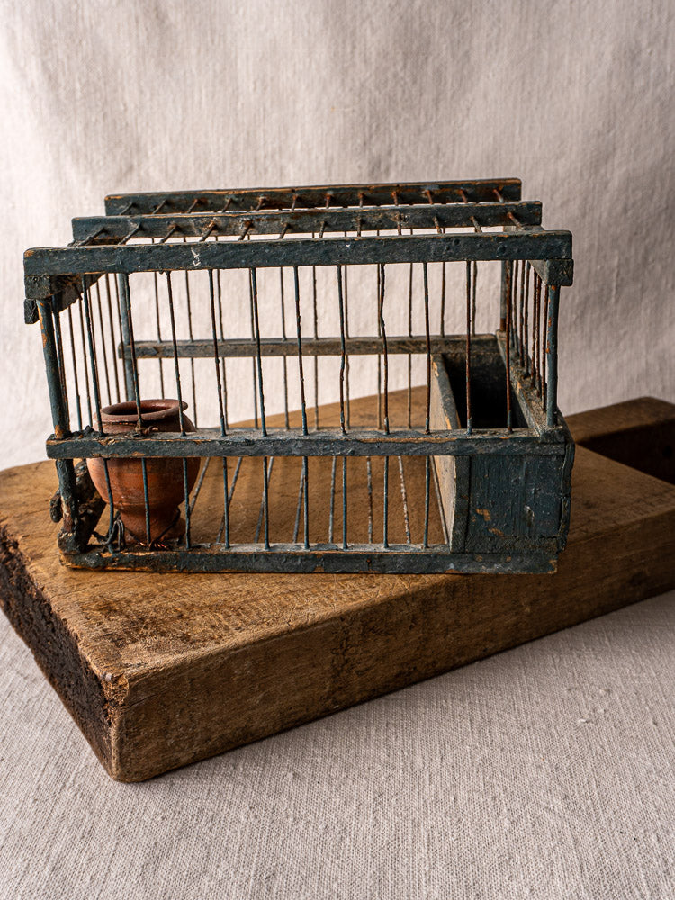 Late 19th-century Provençal bird cage with original blue paint and terracotta cup on wooden boards, neutral fabric backdrop.