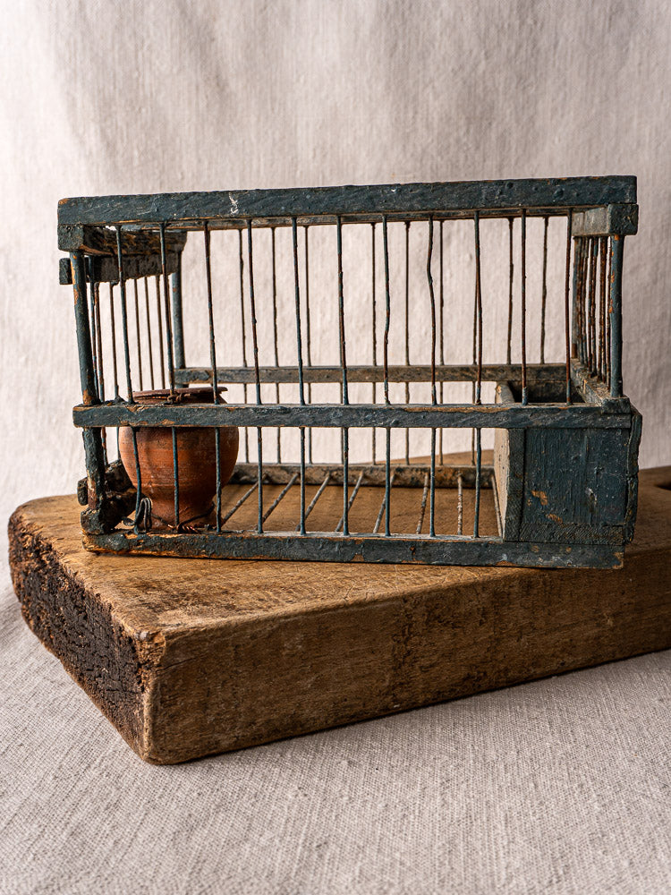 Antique Provençal bird cage with original blue paint and terracotta cup on wooden block, neutral fabric backdrop.