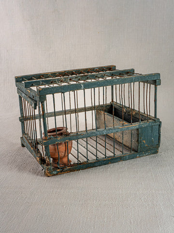 Antique Provençal bird cage with original blue paint, terracotta cup, and metal feeder on a light textured surface.