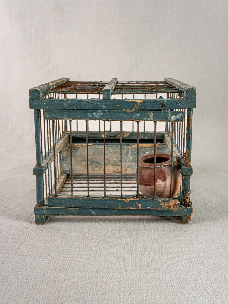 Late 19th-century Provençal bird cage with original blue paint and weathered finish, terracotta cup inside, on neutral fabric.