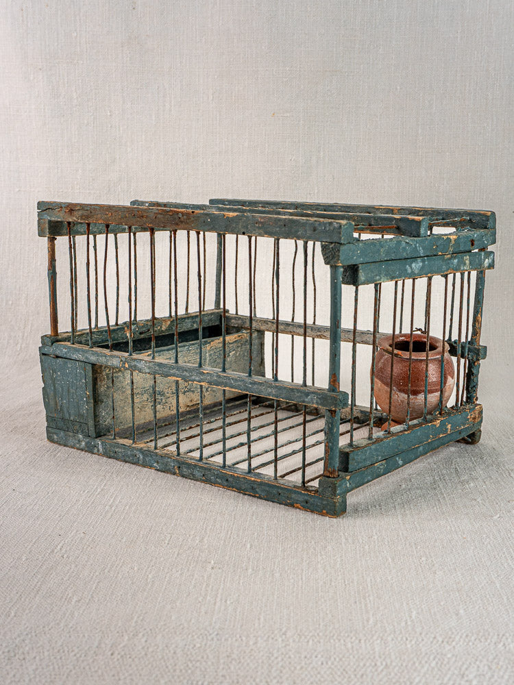 Antique Provençal bird cage with original blue paint, weathered wood, metal bars, and terracotta cup on neutral fabric.