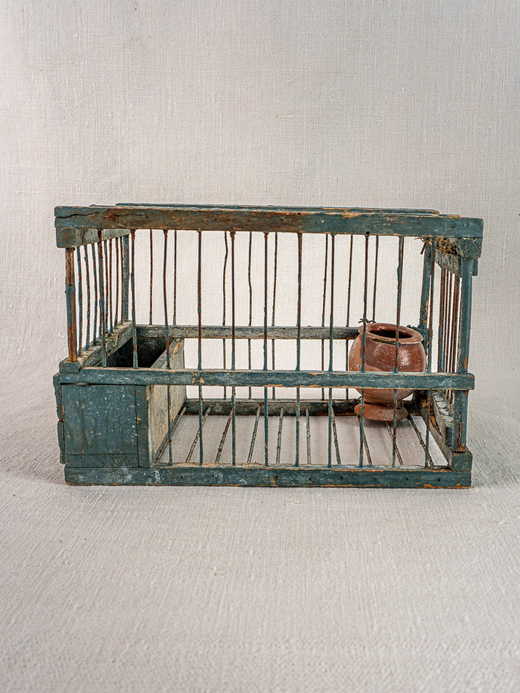Antique Provençal bird cage with original blue paint and terracotta cup on a light textured surface.