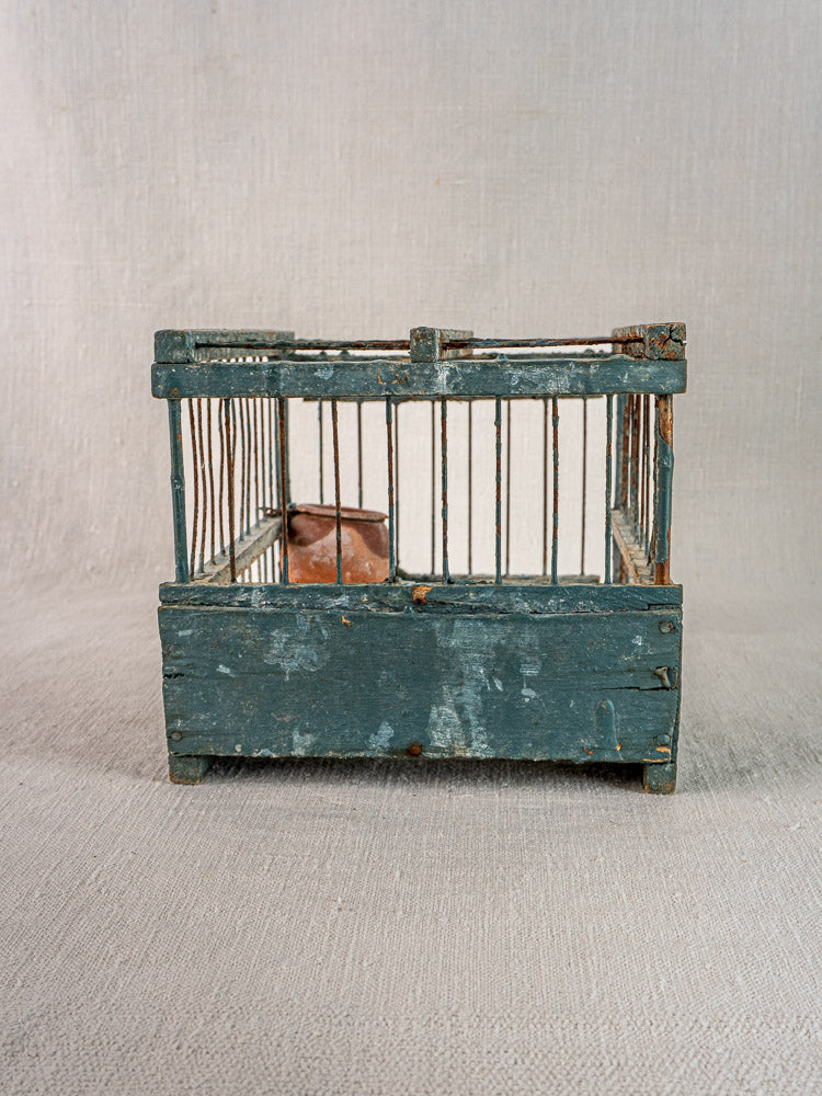Late 19th-century Provençal bird cage with original blue paint and terracotta cup on textured gray surface.