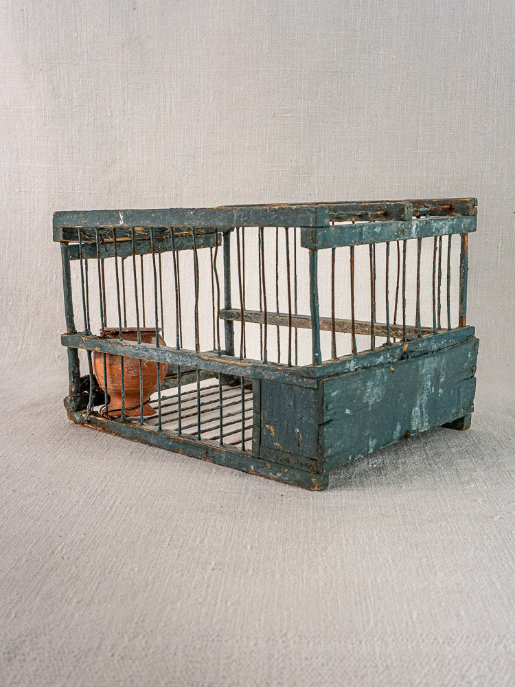 Provençal antique bird cage with original blue paint and terracotta cup, displayed on neutral fabric.