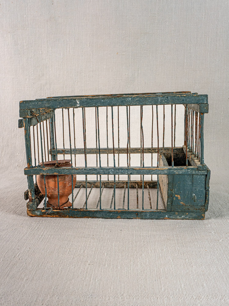 Antique Provençal rectangular bird cage with original blue paint, terracotta cup, and metal bars on neutral fabric background.