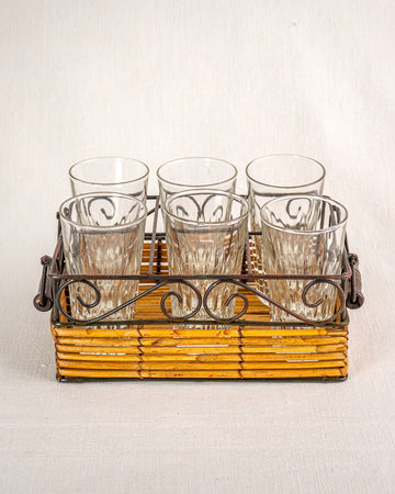 Set of six fluted tumblers in a vintage French bistro glass caddy with wrought iron and rattan on neutral fabric.