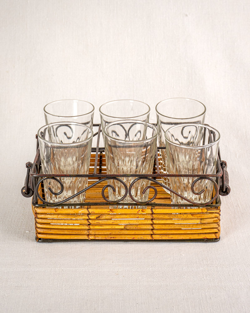 Set of six fluted tumblers in a vintage French bistro glass caddy with wrought iron and rattan on neutral fabric.