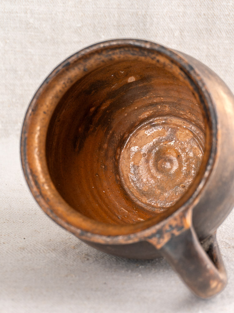 Antique French Provincial glazed earthenware jug with worn, textured interior and single handle on light fabric background.