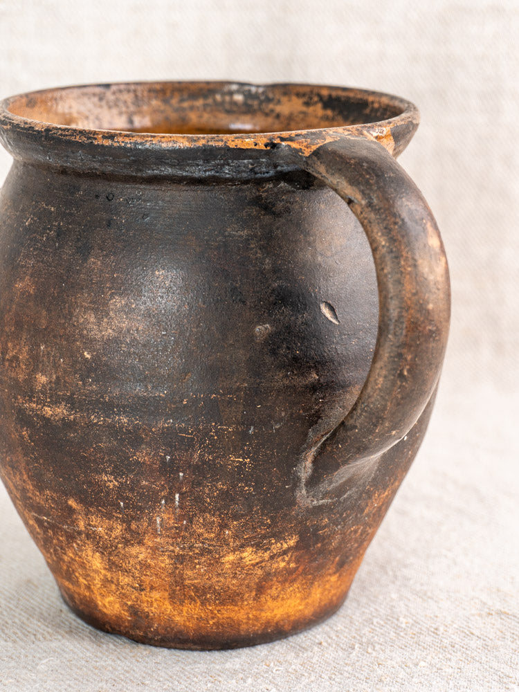 Antique French Provincial glazed earthenware jug with dark brown finish, single handle, and worn texture on light fabric.