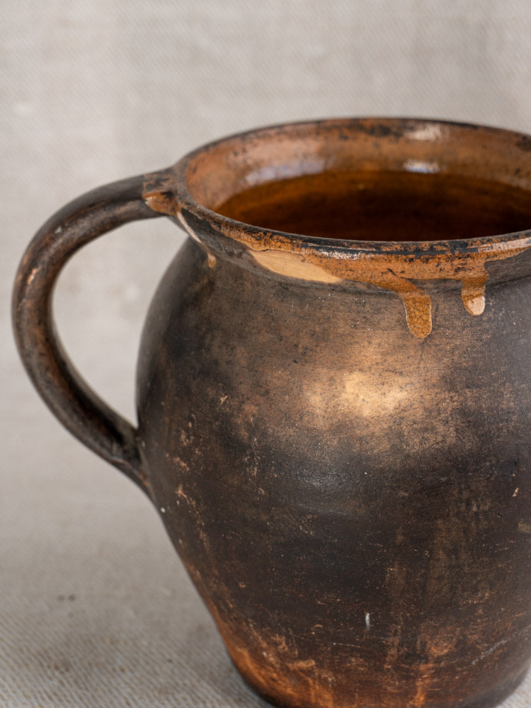 Antique French Provincial earthenware jug with dark brown glaze, single handle, and worn texture on neutral fabric.