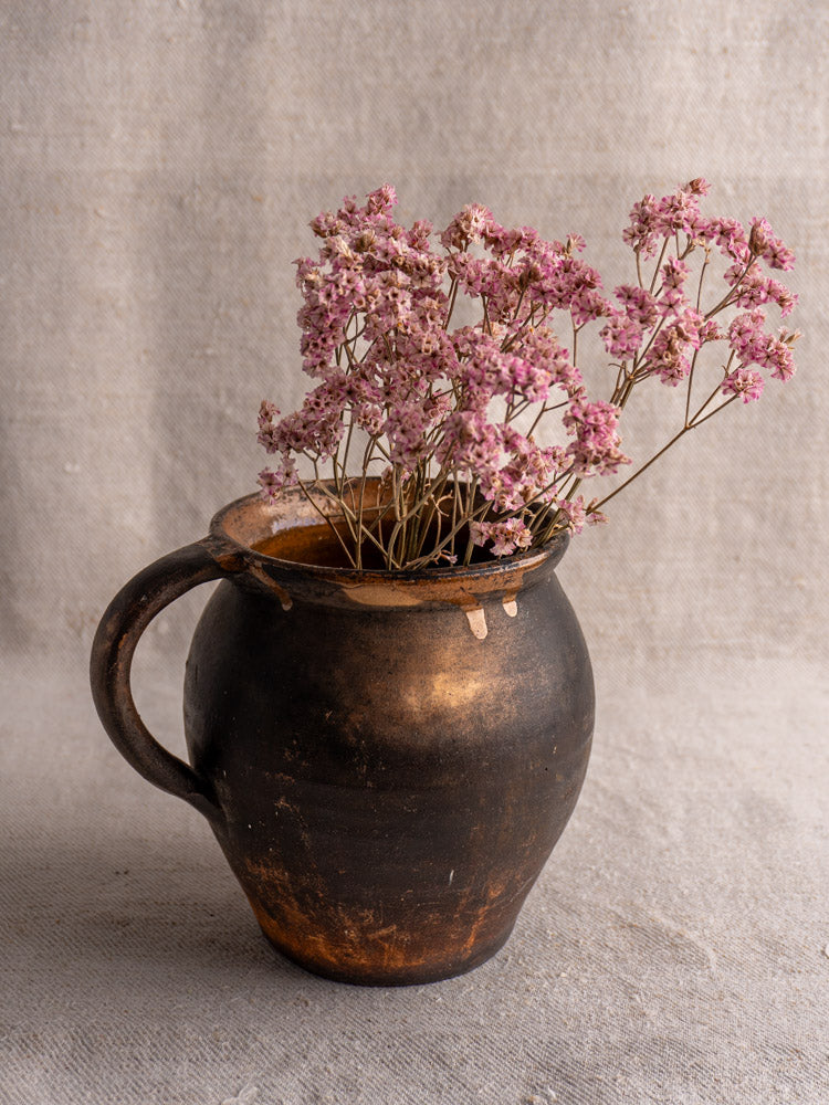 Antique French Provincial glazed earthenware jug with rustic dark brown finish, filled with dried pink flowers on fabric backdrop.