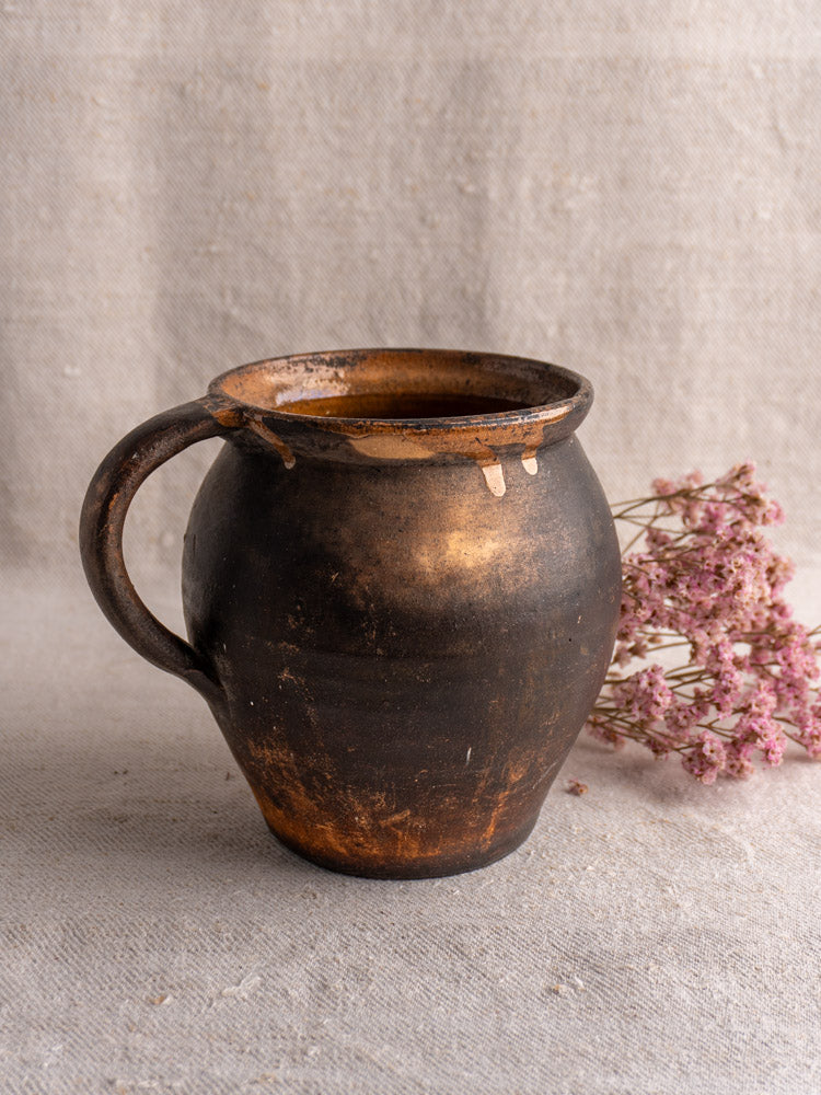 19th-century French Provincial glazed earthenware jug, dark brown with handle and patina, beside dried pink flowers.