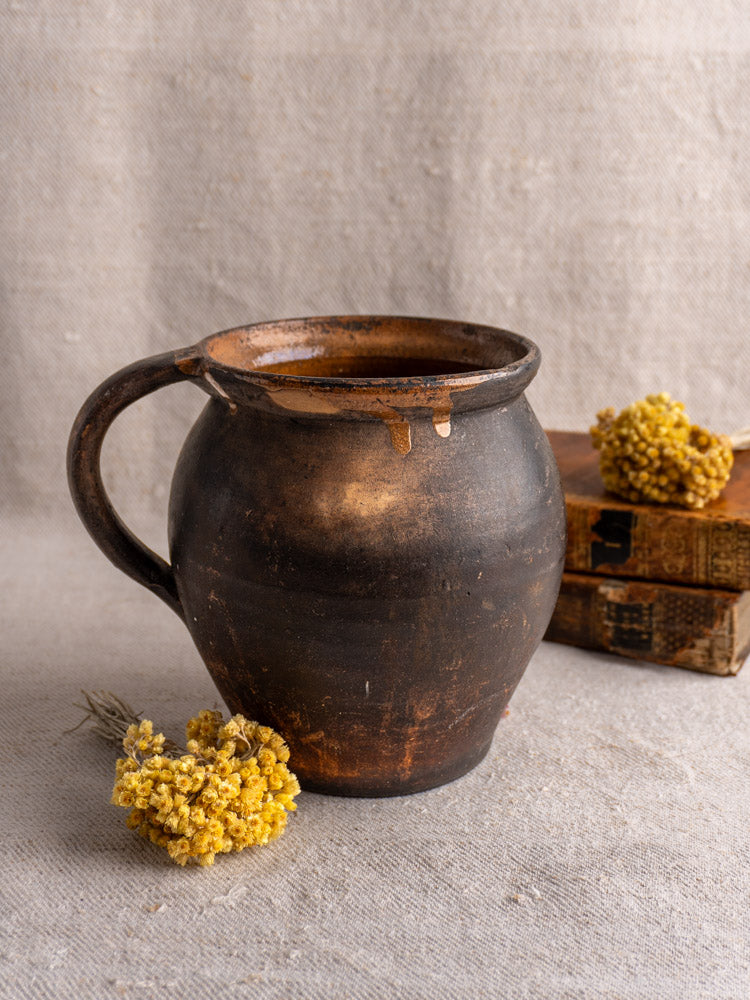 Antique French Provincial glazed earthenware jug with handle on beige cloth, yellow dried flowers, and vintage books behind.