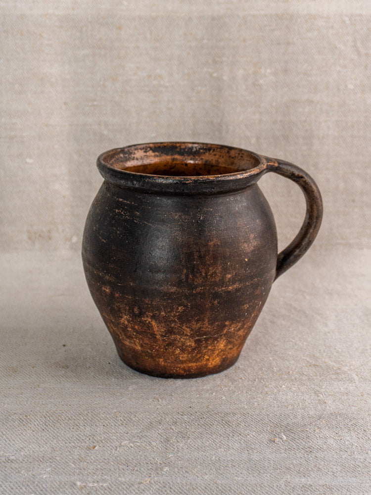 Antique French Provincial glazed earthenware jug with round body and aged handle on textured light fabric.