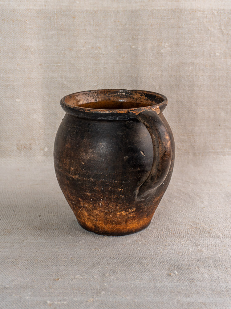 Antique French Provincial glazed earthenware jug with single handle and dark brown glaze on textured fabric surface.