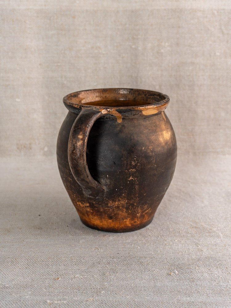 Antique French Provincial glazed earthenware jug with single handle and dark brown, burnished, weathered finish on beige surface.