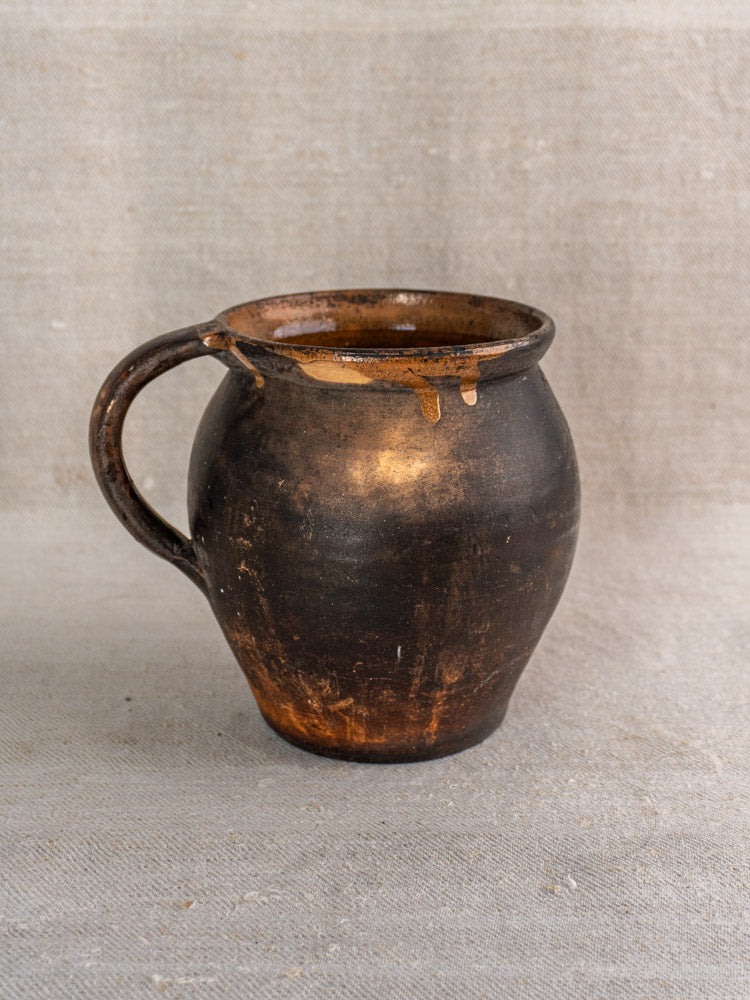 Antique 19th-century French earthenware jug with rounded body, worn handle, and glaze drips on neutral textured fabric.