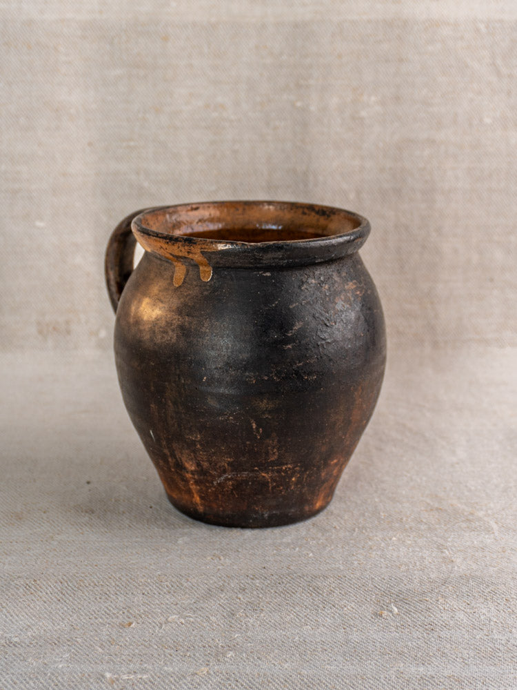 19th-century French Provincial earthenware jug with dark brown glaze, round body, wide mouth; on textured beige fabric.