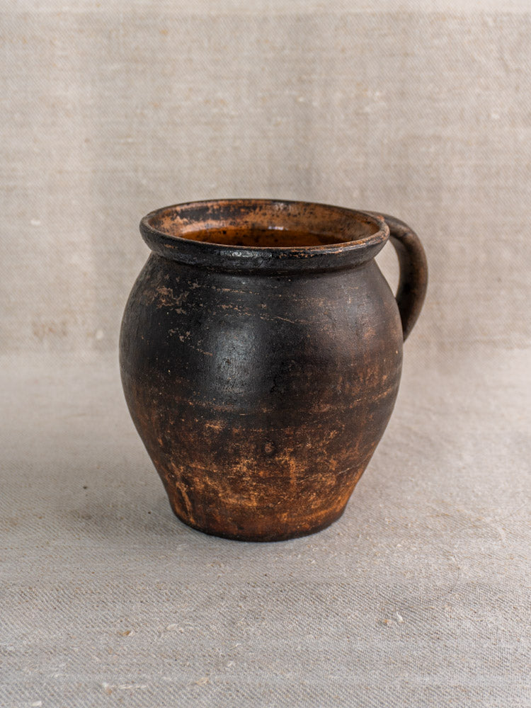 19th-century French Provincial earthenware jug with dark brown glaze and single handle on beige textured fabric.