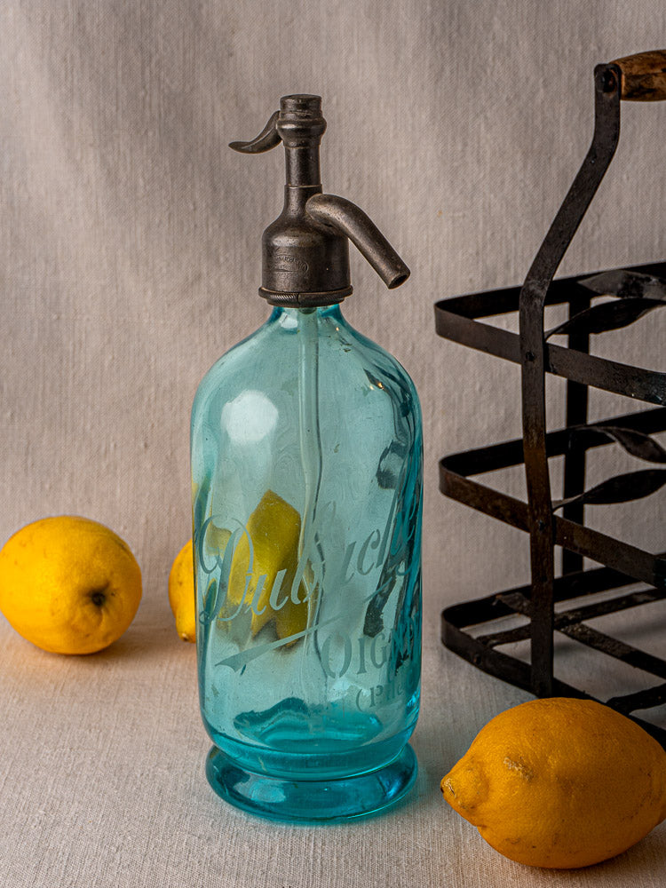 Antique French blue glass soda siphon with metal spout beside black crate and lemons.