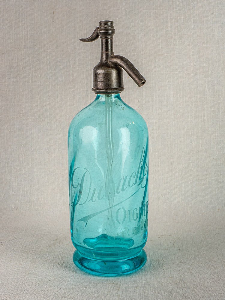Antique French J. Dubuche blue glass seltzer bottle with metal siphon on neutral surface.
