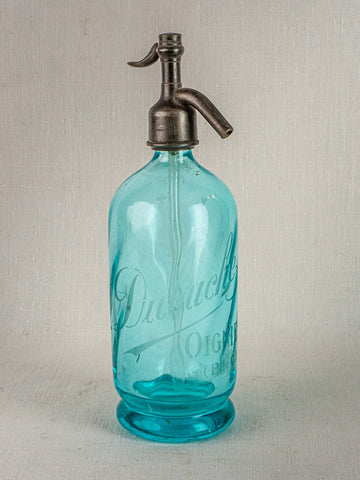 Antique French J. Dubuche blue glass seltzer bottle with metal siphon on neutral surface.