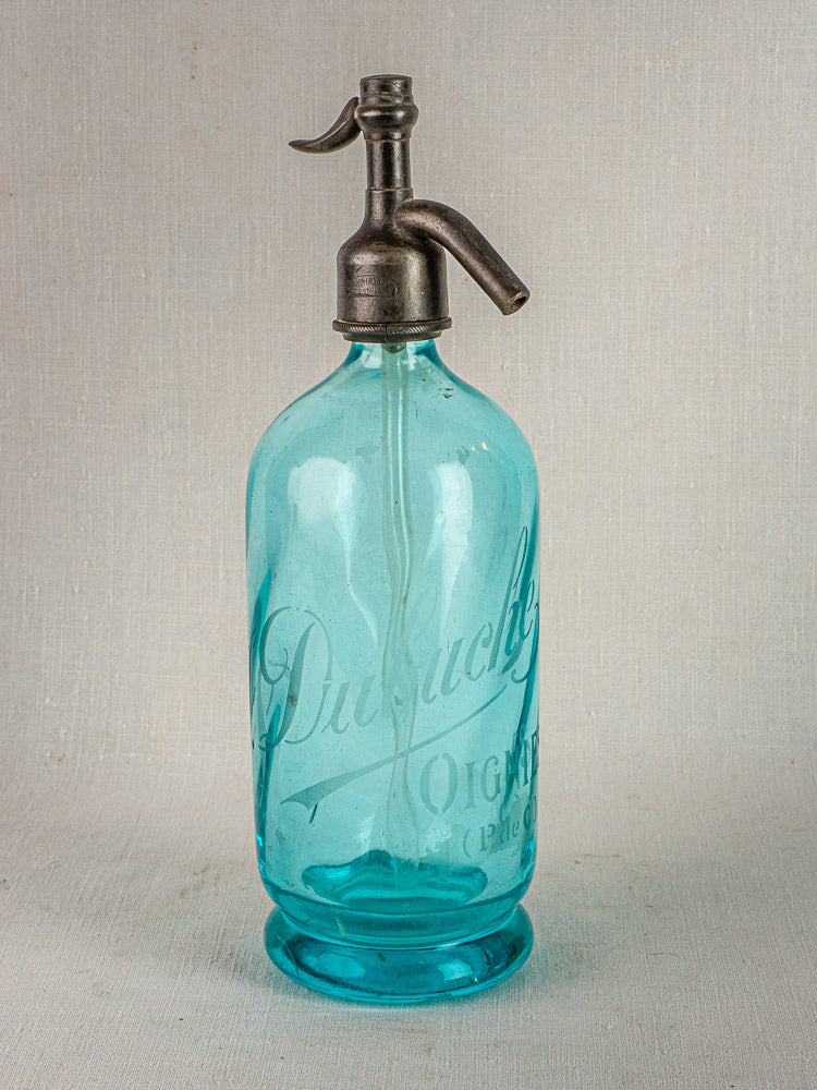 Antique French J. Dubuche blue glass seltzer bottle with metal siphon on neutral surface.