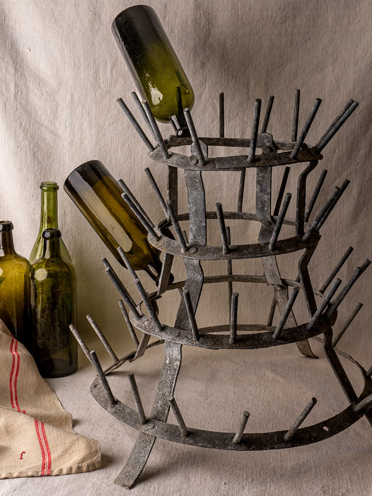 Antique French galvanized zinc bottle dryer with prongs, two green bottles, and striped cloth on neutral background.