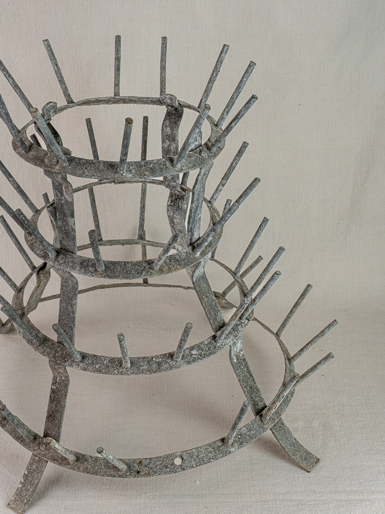 Antique French bottle dryer circa 1900 in galvanized zinc with four circular tiers and pegs against a neutral fabric backdrop.