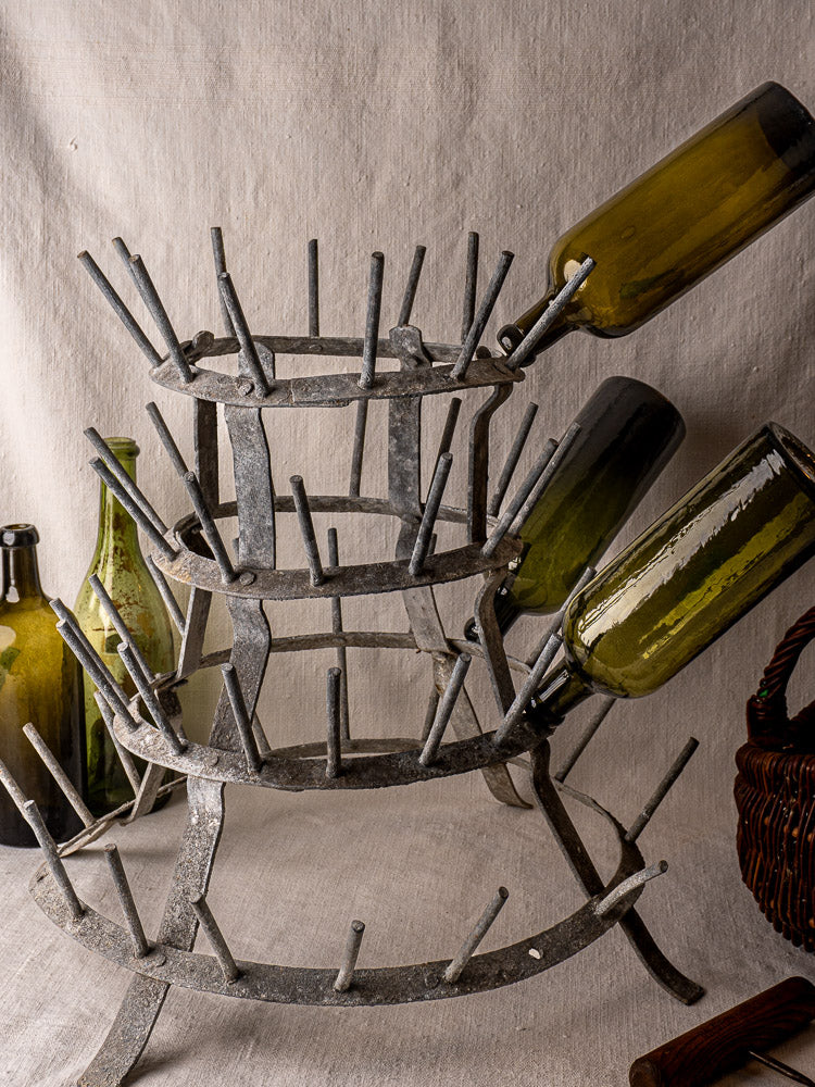 Antique French galvanized zinc bottle dryer with green glass bottles and woven basket on beige fabric backdrop.