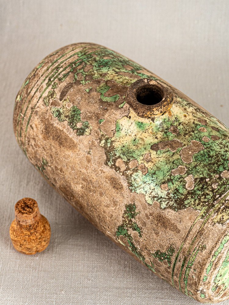 Antique French terracotta bouillotte with weathered green patina and small opening, beside cork stopper on neutral fabric.
