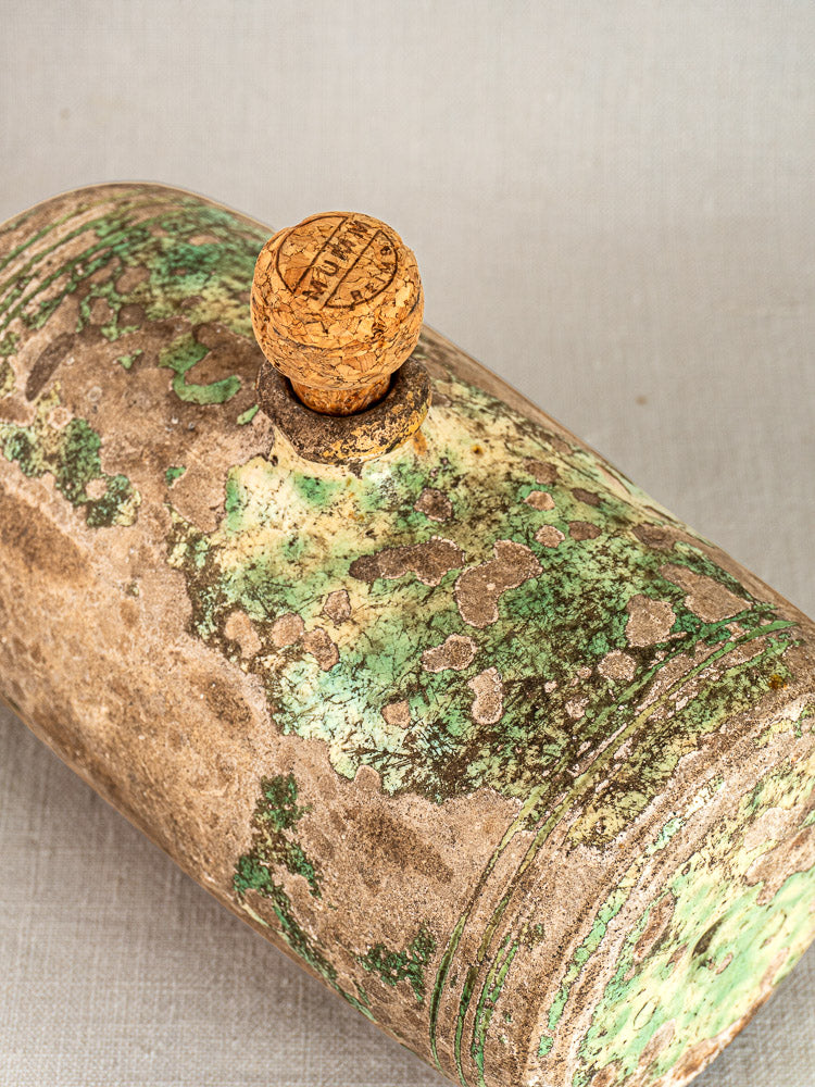 Weathered glass bottle with cork marked HOLLAND beside 18th-century French terracotta Bouillotte on pale fabric.