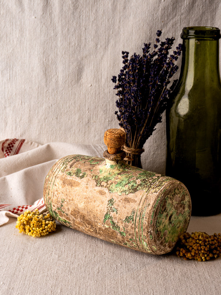 Antique green butter churn with wooden handle beside terracotta Bouillotte, green glass bottle, and linen on beige fabric.